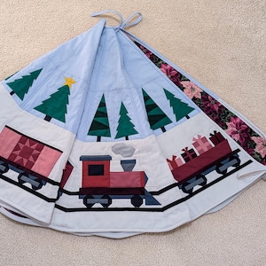 Op de afbeelding: Een ronde, doorgestikte kerstboomrok met een lichtblauwe achtergrond, groene dennenbomen en een gele ster. De onderste rand toont een trein met een rode locomotief, een roze goederenwagon en een wagon met cadeautjes op zwarte rails.