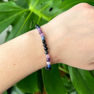 Könnte beinhalten: Ein mehrfarbiges Perlenarmband mit schwarzen Lavasteinen, rosa, lila und blauen Perlen. Das Armband befindet sich am Handgelenk einer Person.
