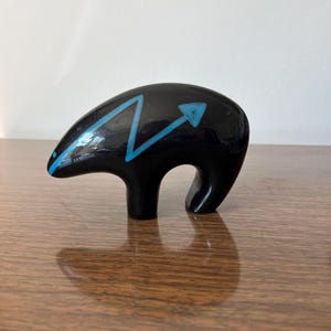 May include: A glossy black bear figurine with a blue arrow design. The bear has a rounded body and a curved leg, standing on a wooden surface. The blue arrow design is on the bear's back.