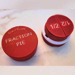 Könnte beinhalten: Rotes Plastik-Bruchkuchen-Set mit den Worten "Love 2 Learn Fraction Pie" auf dem Deckel und den Brüchen "1/2" und "1/2" auf den Stücken.