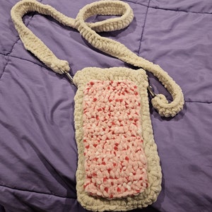 May include: A rectangular crochet crossbody bag made of thick, cream-colored yarn. The center panel features a textured pink and white speckled pattern. The bag includes a long, matching cream-colored crochet strap attached with silver metal clips.