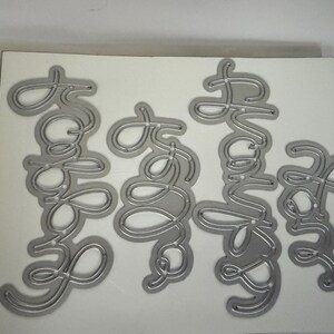 May include: Silver metal die cuts spelling out "Hello", "Baby", "Thank You", and "Yay" in a cursive font. The dies are on a white background. The words are connected and have a delicate, elegant appearance.