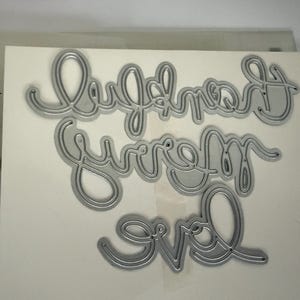 May include: Silver metal die cuts spelling out "Love, Forever, Ever" in a cursive font. The dies are on a white background with the text "EXPRESSIONS THINLITS DIES (132171)" on the left side.