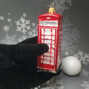 May include: A red glass ornament shaped like a British telephone booth, with the word "TELEPHONE" across the top. The ornament has silver glitter accents and a gold-colored metal loop for hanging. A white sphere sits next to the ornament.