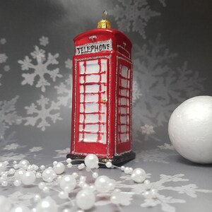 May include: A red Christmas ornament shaped like a British telephone booth, with the word "TELEPHONE" across the top. The ornament has white window panes and silver glitter accents. A white pearl garland and a white sphere are in the foreground.