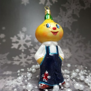 May include: A whimsical Christmas ornament shaped like a smiling onion character. The ornament features a yellow onion head, blue eyes, and a red nose and mouth. It wears a white shirt with a blue overall dress. The ornament is decorated with glitter.