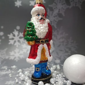 Puede incluir: Adorno navideño con forma de Papá Noel. El adorno es rojo, blanco y azul, con un cinturón negro y hebilla dorada. Papá Noel sostiene un árbol de Navidad verde. El adorno mide aproximadamente 13 cm de alto.