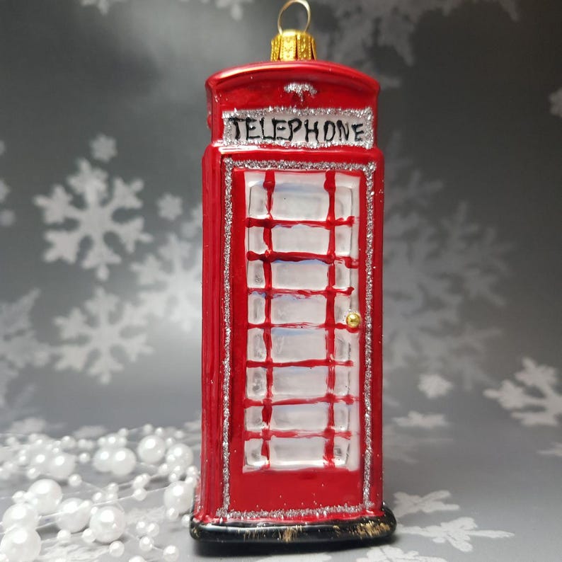 May include: A red Christmas ornament shaped like a British telephone booth. The ornament has a gold loop for hanging and is trimmed with silver glitter. The word "TELEPHONE" is written across the top. The door has a gold knob.