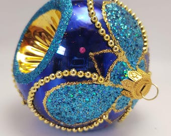 May include: A blue Christmas ornament with gold and turquoise glitter accents. The ornament has a gold cap and a loop for hanging. The design includes gold beaded trim and turquoise glitter patterns.