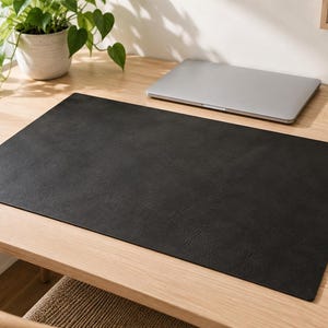 Könnte beinhalten: Eine rechteckige, schwarze Schreibtischunterlage mit matter Oberfläche und geraden Kanten. Die Unterlage weist eine dezente, feinkörnige Textur auf und bietet eine flache, gleichmäßige Arbeitsfläche für einen Computer oder zum Schreiben.