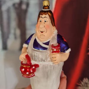 以下が含まれることがあります： 厳しい表情のキャラクターを描いたガラス製のクリスマスオーナメント。白いエプロンに青と白の衣装を着て、赤いティーポットと白い水玉模様の赤い容器を持っています。オーナメントは金の王冠をしています。