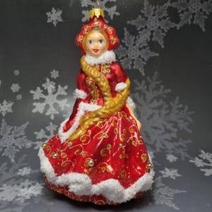 May include: A red glass Christmas ornament depicting a woman in a red dress with a white fur trim and a gold braid. She is wearing a red hat with gold accents and has a white fur collar. The ornament is decorated with gold glitter and red beads.