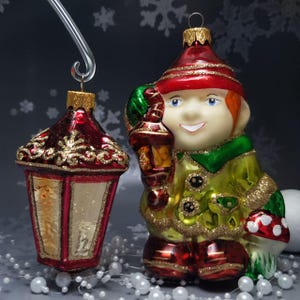 Könnte beinhalten: Zwei Weihnachtsschmuckstücke: eine rot-goldene Laterne und ein lächelnder Wichtel mit einer Laterne. Der Wichtel trägt einen roten Hut, einen grünen und goldenen Mantel und rote Stiefel. Beide Ornamente haben goldene Glitzerakzente.