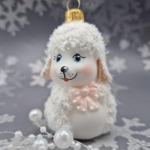 May include: A white poodle-shaped Christmas ornament with a textured, fuzzy finish. The dog has blue eyes, a black nose, and a pink bow at the neck. It features a gold-coloured metal hanging loop at the top and a smooth, white lower body.