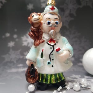May include: A whimsical Christmas ornament depicting a doctor figure with a monkey on his shoulder. The doctor wears a white coat, green and black striped pants, and a red cross hat. The ornament has a gold-colored top.