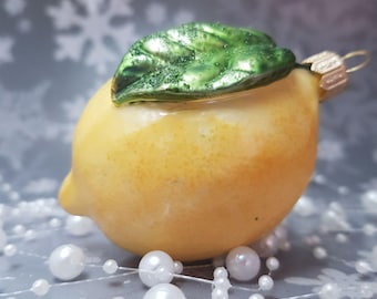 Op de afbeelding: Een citroenvormige kerstversiering met een groen blad en een goudkleurige metalen lus. De versiering is geel met een glanzende afwerking. Witte parel kralen staan op de voorgrond.