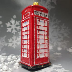 May include: A red telephone booth Christmas ornament with silver glitter trim. The ornament has the word "TELEPHONE" on top. The windows are white with a grid pattern. A gold-colored loop is at the top for hanging.