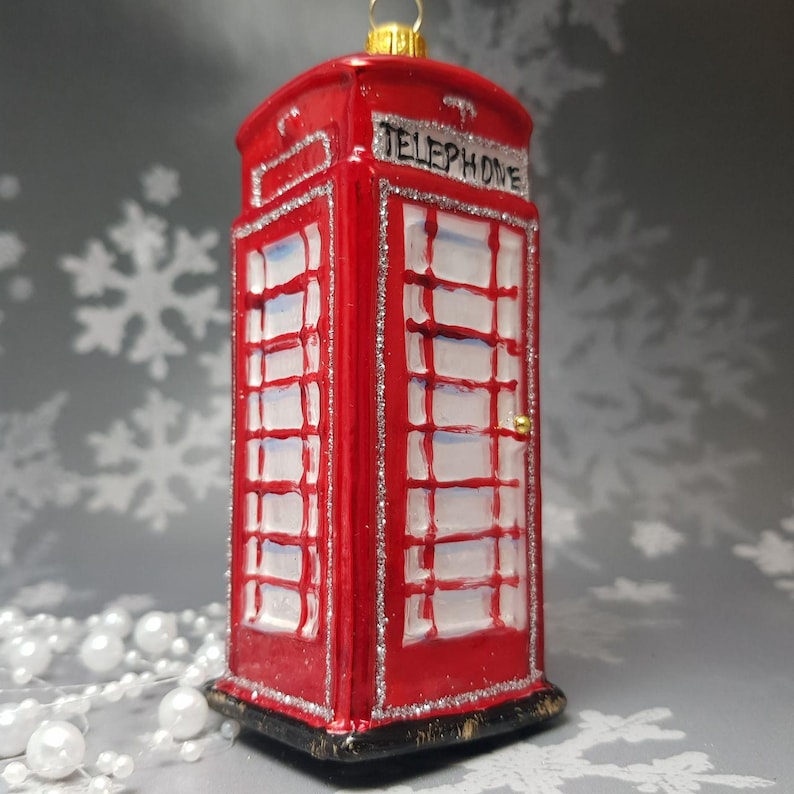 May include: A red telephone booth Christmas ornament with silver glitter trim. The ornament has the word "TELEPHONE" on top. The windows are white with a grid pattern. A gold-colored loop is at the top for hanging.