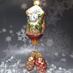 May include: A detailed Christmas ornament shaped like a cuckoo clock, with a gold and red finish. The clock face is white with black numerals. Two red and gold pinecone ornaments hang below. Festive holiday decor.