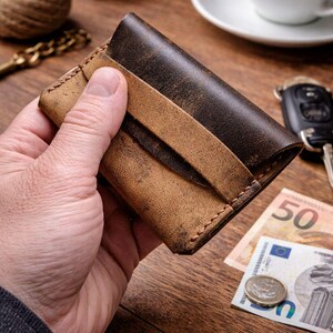 May include: A brown leather wallet with a distressed finish, held in a hand. The wallet features a strap closure and visible stitching. The background includes a cup of coffee, keys, and currency. The wallet has a vintage appearance.