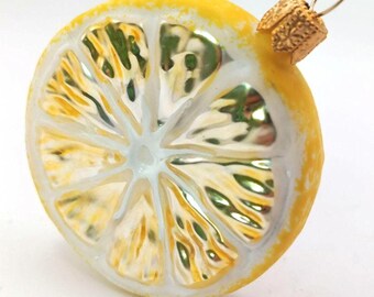 Op de afbeelding: Een glazen kerstornament in de vorm van een citroen schijf. De gele schil en de witte segmenten zijn gedetailleerd. Het heeft een goudkleurige metalen dop en lus om op te hangen. Een feestelijke decoratie.