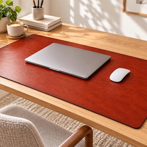 Op de afbeelding: Een rechthoekige, donkerroodbruine bureauonderlegger met een gladde, leerachtige textuur. De mat ligt op een houten oppervlak en biedt plaats aan een gesloten zilverkleurige laptop en een witte computermuis voor een opgeruimd bureau.