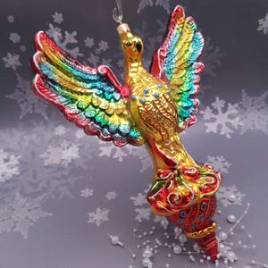 Może przedstawiać: Kolorowa szklana ozdoba choinkowa w kształcie ptaka z rozłożonymi skrzydłami. Ptak ma złote, czerwone, niebieskie i zielone odcienie. Ozdoba jest zawieszona na srebrnym sznurku.