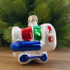 May include: A vintage Christmas tree ornament in the shape of a train. It has a white body with red, green, and blue details. The ornament has a gold-coloured metal cap and is set against a blurred green background.