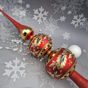 May include: A red and gold glass Christmas tree topper featuring two spherical ornaments decorated with hand-painted gold floral patterns, glitter accents, and a pointed finial top. The piece has a shiny finish with black dots.