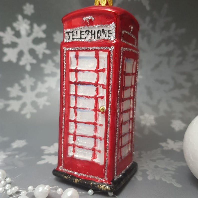 May include: A red Christmas ornament shaped like a British telephone booth. The ornament has the word "TELEPHONE" at the top and is trimmed with silver glitter. The door has a gold-colored knob and white panes.