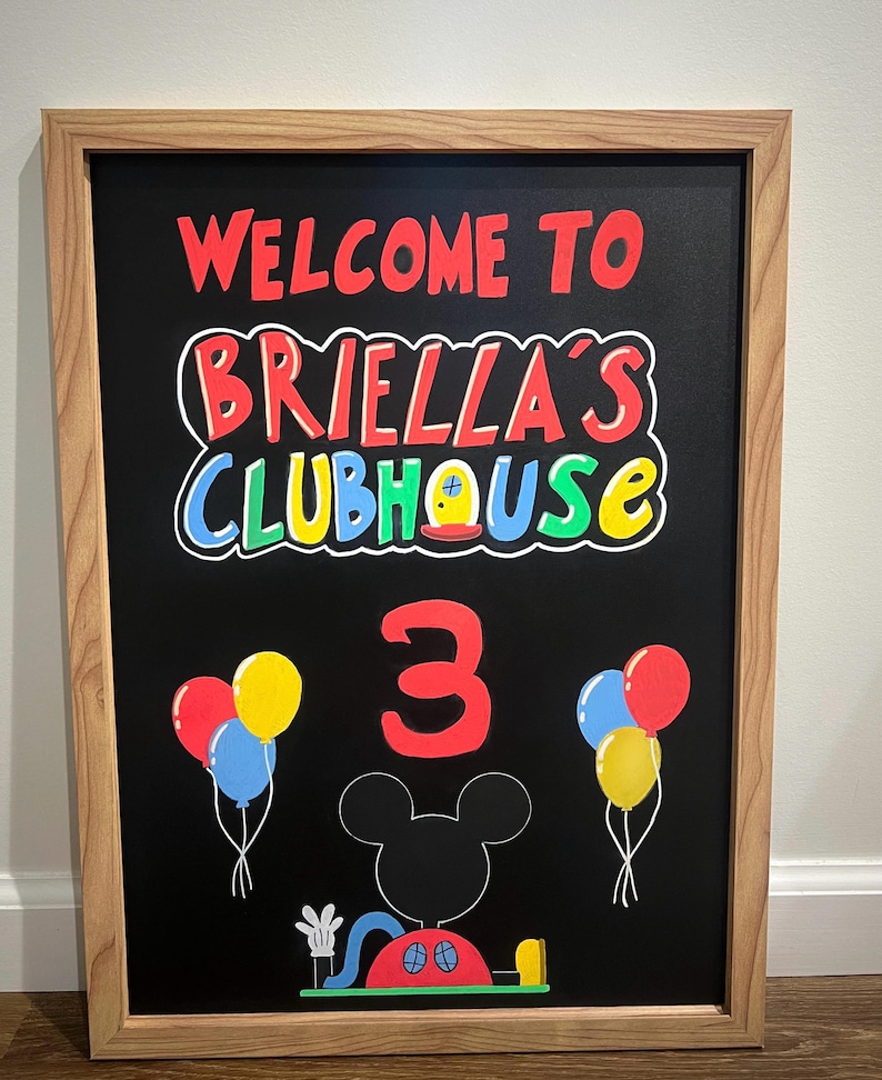 Pode incluir: Um quadro-negro emoldurado com o texto "WELCOME TO BRIELLA'S CLUBHOUSE" em vermelho, branco, azul e amarelo. O n&uacute;mero "3" &eacute; vermelho, com Mickey Mouse e bal&otilde;es. A moldura &eacute; marrom claro.