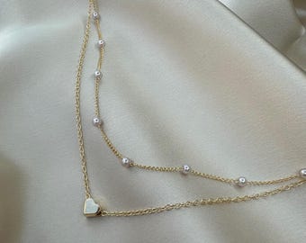May include: A delicate gold-colored necklace with two strands. The top strand features small, evenly spaced pearls, while the bottom strand has a small heart-shaped pendant. The necklace is displayed on a satin-like, cream-colored surface.