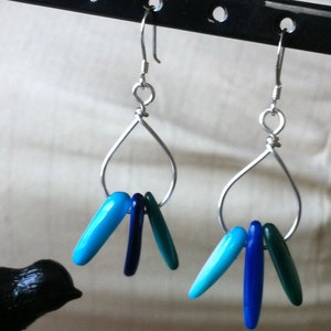 May include: A pair of silver-toned teardrop earrings. Each earring features a silver wire teardrop shape with three dangling, elongated beads in shades of blue and green. The earrings hang from a black display stand.