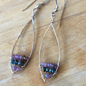 May include: A pair of silver teardrop earrings. Each earring features a wire frame with three rows of small, colorful gemstones in shades of purple, blue, and green. The earrings have a simple, elegant design.