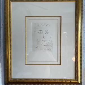 May include: A framed line drawing of a human face in black ink on a white background. The artwork is centered within a cream-colored mat and enclosed in a rectangular gold-toned frame with a textured, beaded border detail along the inner edge.