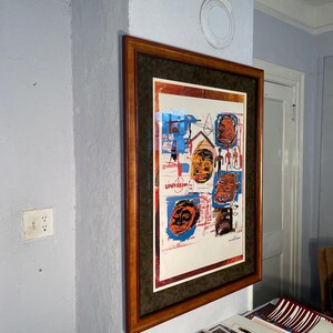 K&ouml;nnte beinhalten: Gerahmtes Kunstwerk mit farbenfrohen abstrakten Gesichtern und Figuren. Das Kunstwerk befindet sich in einem braunen Holzrahmen mit einem dunkelgr&uuml;nen Passepartout. Das Kunstwerk ist an einer hellblauen Wand befestigt.