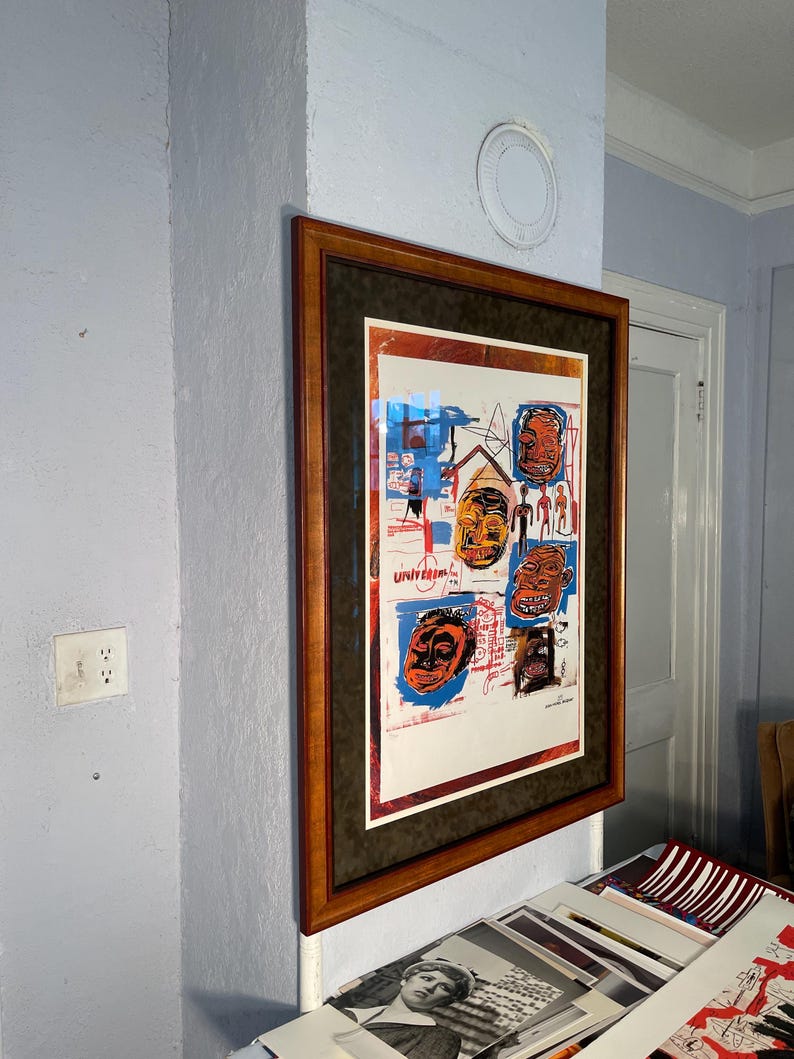 K&ouml;nnte beinhalten: Gerahmtes Kunstwerk mit farbenfrohen abstrakten Gesichtern und Figuren. Das Kunstwerk befindet sich in einem braunen Holzrahmen mit einem dunkelgr&uuml;nen Passepartout. Das Kunstwerk ist an einer hellblauen Wand befestigt.