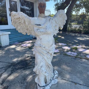 May include: A life-sized, weathered white statue of a winged figure with a human head and torso. The figure features textured, outstretched wings, a draped garment, and a dark-toned face. A metal chain is wrapped around the base of the statue.