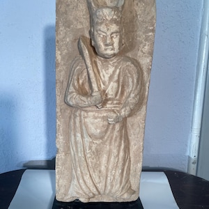 Puede incluir: Escultura en relieve beige de una figura sosteniendo una espada. La figura lleva una túnica y un tocado alto. La escultura está montada sobre una base negra. Forma rectangular.