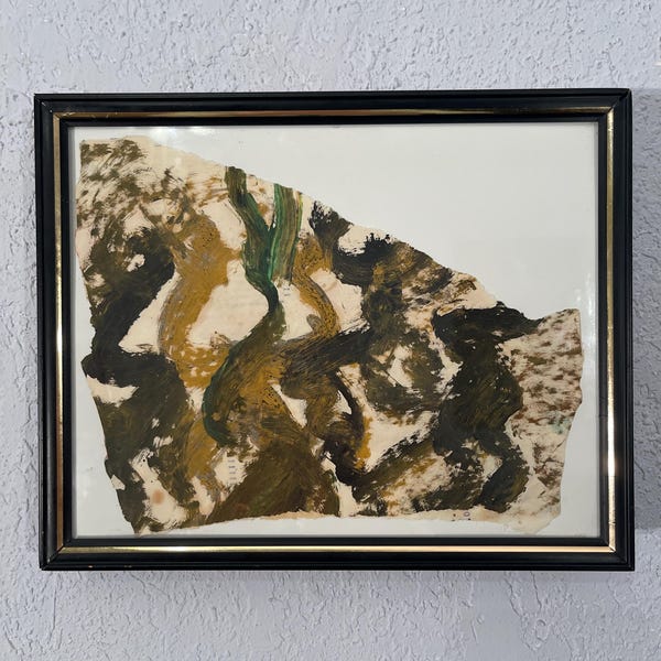 May include: Abstract artwork in a black frame. The art piece features a torn paper with brushstrokes in shades of brown, green, and black. The frame has a gold inner edge, and the background is a textured, light grey wall.