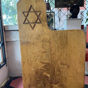 May include: Wooden chair with a Star of David cutout on the back. The chair has a natural wood grain finish and a simple, traditional design. The chair is likely used for religious or ceremonial purposes.