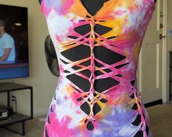 Może przedstawiać: Sukienka bez rękawów z wzorem tie-dye w odcieniach różu, pomarańczu i fioletu. Odzież posiada dekolt w serek oraz centralny pionowy wycięty wzór z plecionymi detalami z tkaniny i małymi metalowymi koralikami wzdłuż przedniej części tułowia.