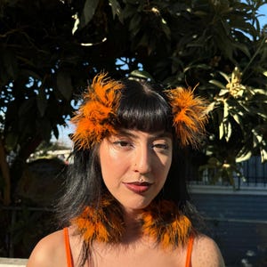 Puede incluir: Una persona lleva un top naranja brillante sin mangas con pendientes y diadema a juego de plumas naranja y negras.