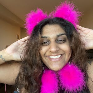 May include: A vibrant pink feather headband with matching ear-shaped details and two large, fluffy feather pendants. The accessories are a bright fuchsia colour, creating a playful and eye-catching look.