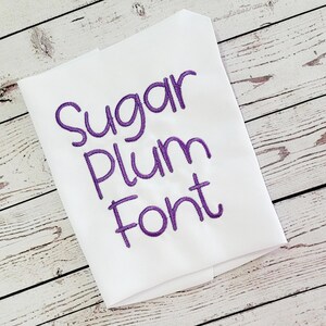 Puede incluir: Tela blanca con las palabras "Sugar Plum Font" bordadas en hilo morado. La tela está doblada y en ángulo, sobre un fondo de vetas de madera clara.