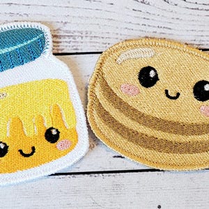 May include: Two embroidered patches, one of a jar of honey and the other of a stack of pancakes. Both patches have cute cartoon faces with smiling mouths and big eyes.