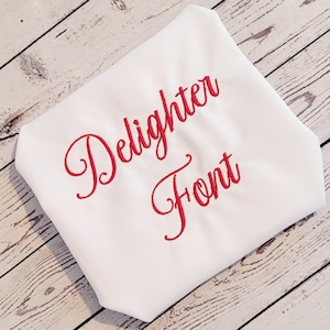 Pode incluir: Tecido branco com as palavras "Delighter Font" bordadas em vermelho com escrita cursiva. O tecido está dobrado e inclinado, sobre um fundo de grão de madeira clara.