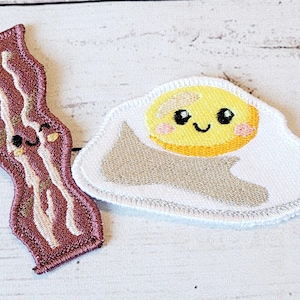 Può includere: Due toppe ricamate, una di una striscia di pancetta sorridente e l'altra di un uovo fritto sorridente. Entrambe le toppe sono bianche con ricami marroni e gialli.