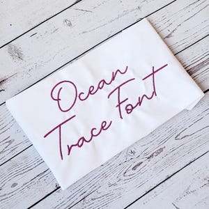 Puede incluir: Tela blanca con el texto "Ocean Trace Font" bordado en rosa.