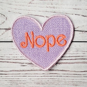 Puede incluir: Parche morado en forma de corazón con la palabra "Nope" bordada en naranja.